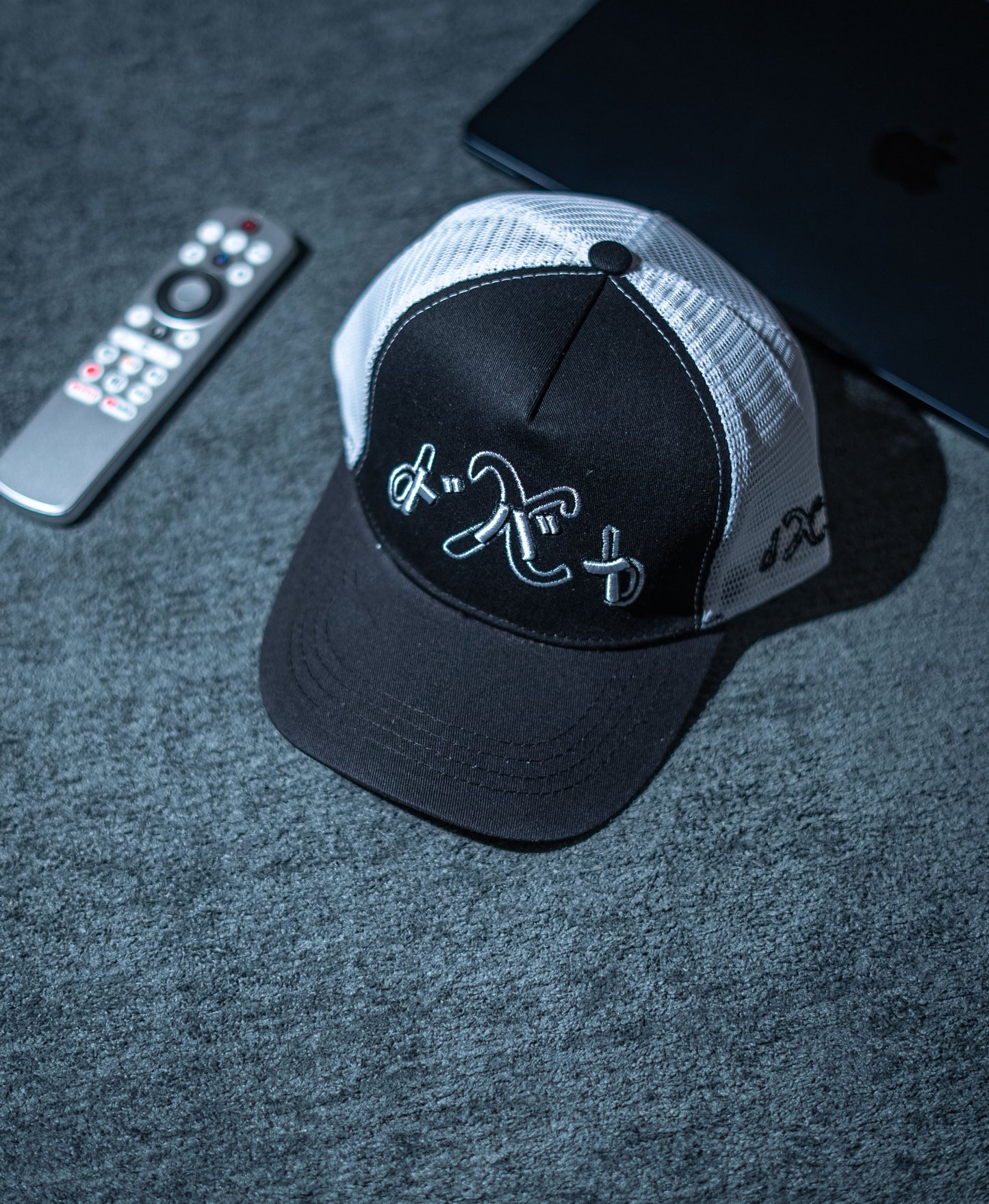 Black and gray cap with embroidered design on a dark surface with remote control and phone in Pakistan by Caps Nation