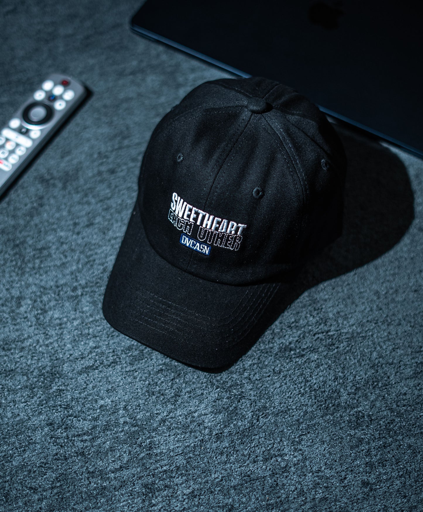 Black cap with text on a dark textured surface with remote control and laptop in Pakistan by Caps Nation