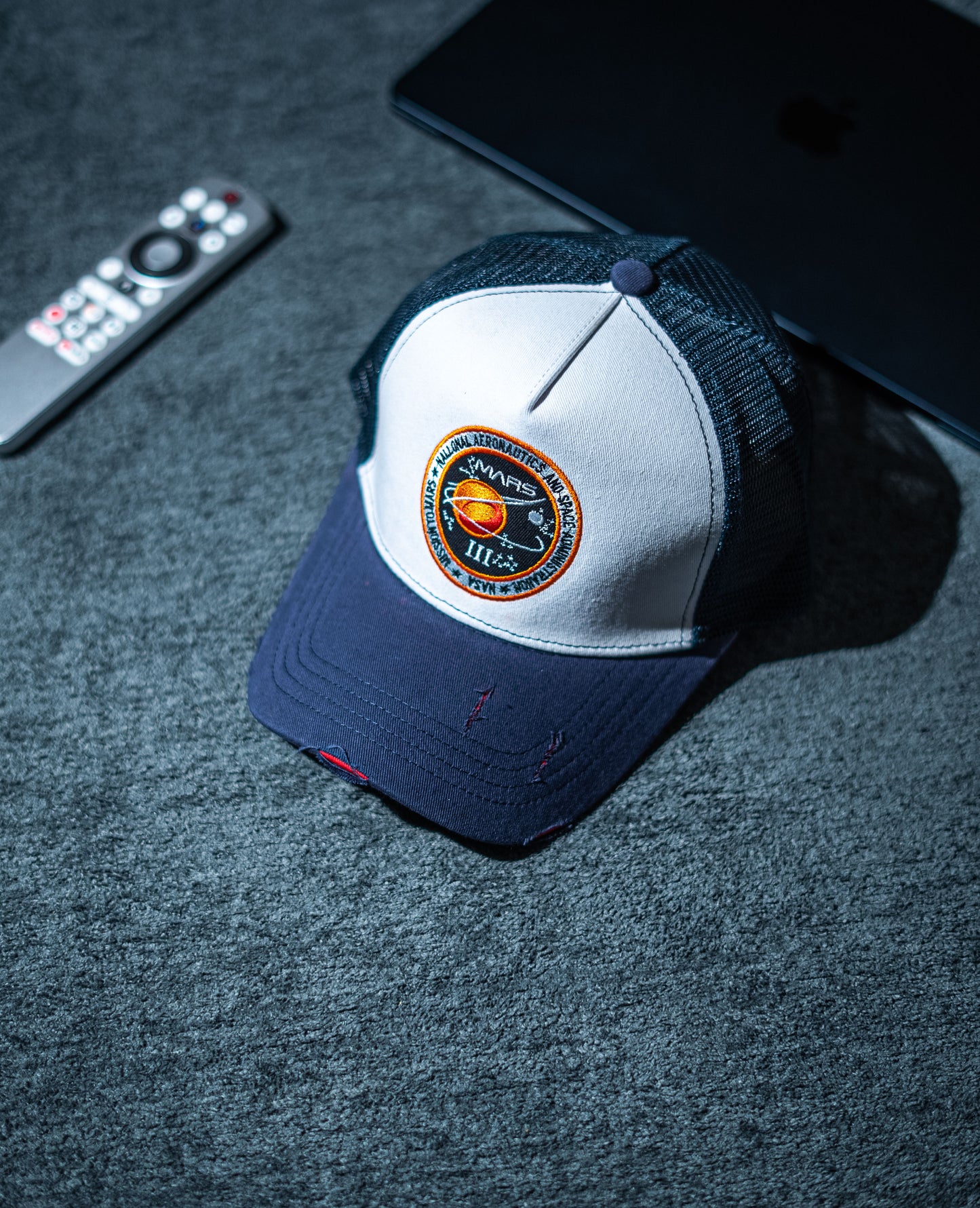 Blue and white cap with a logo on a dark surface next to a remote control and tablet in Pakistan by Caps Nation