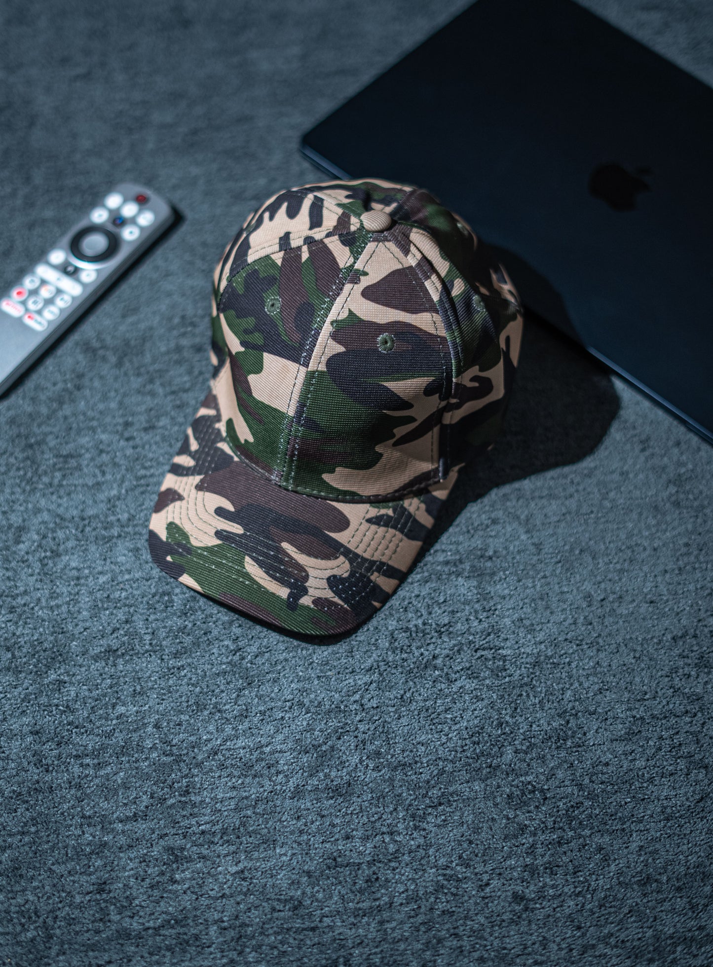 Camouflage baseball cap on a dark textured surface with a remote control and electronic device in Pakistan by Caps Nation