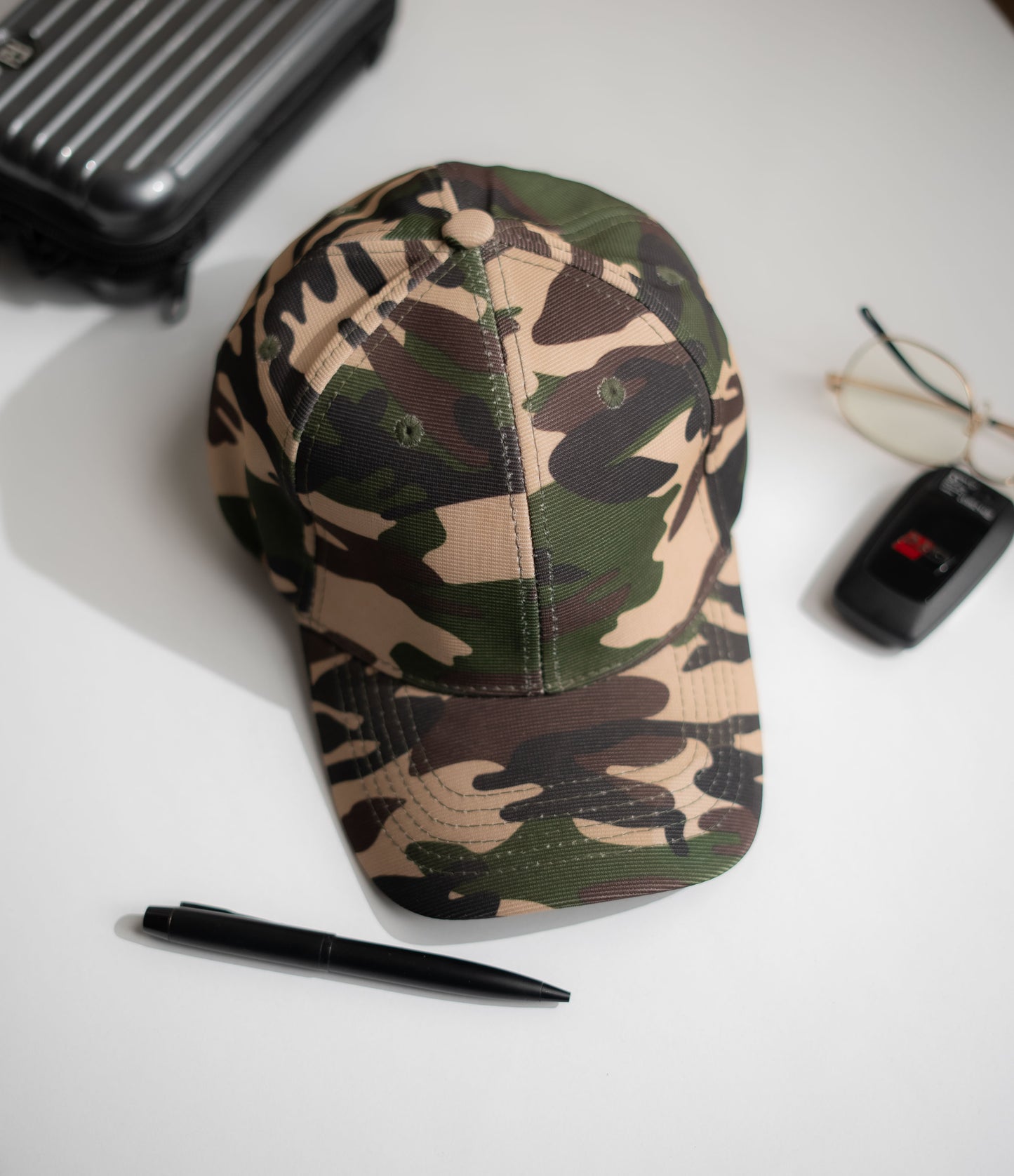 Camouflage baseball cap on a white surface with a pen, glasses, and black case in Pakistan by Caps Nation