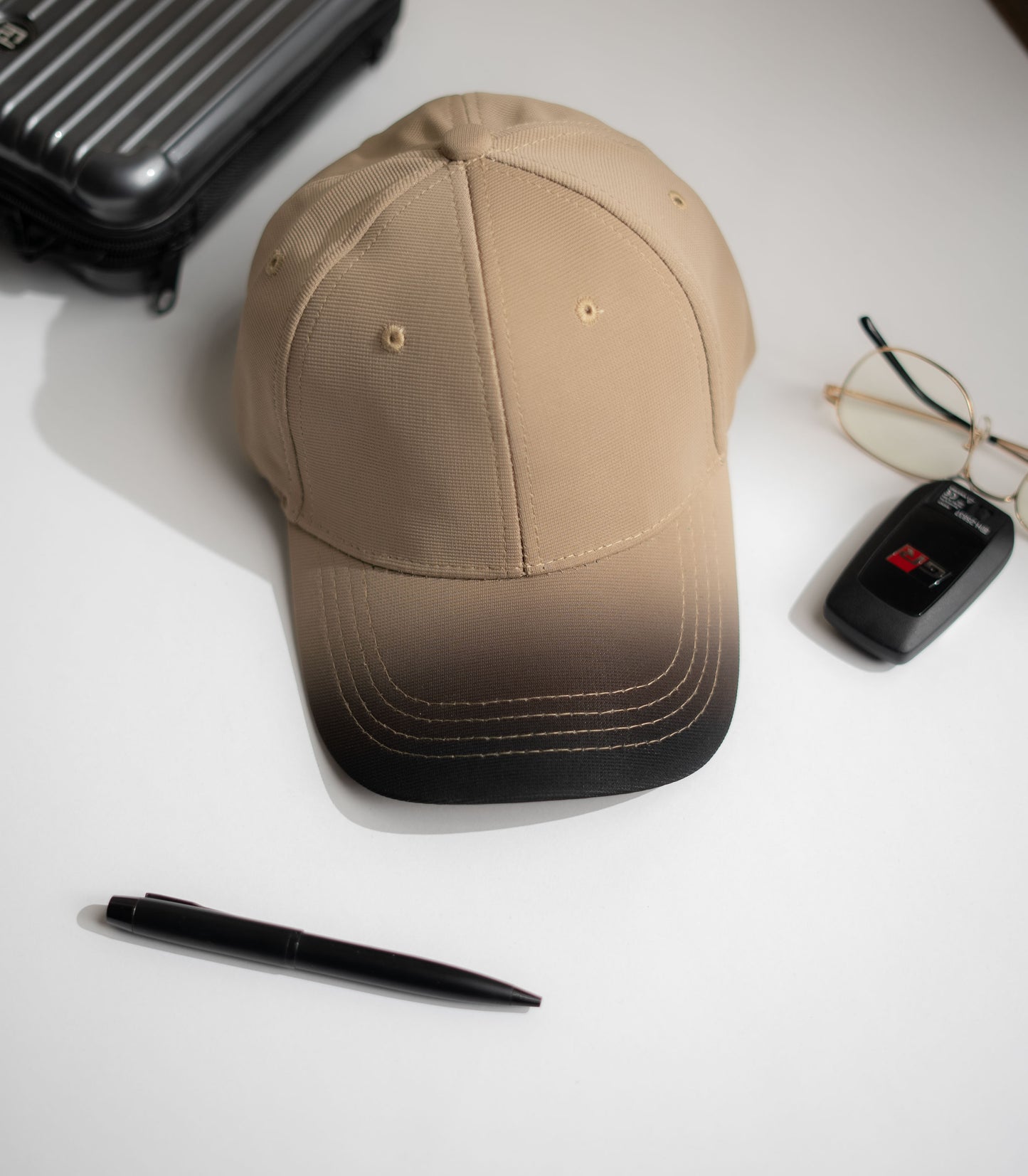 Beige cap with gradient design, pen, glasses, and suitcase on a white surface in Pakistan by Caps Nation
