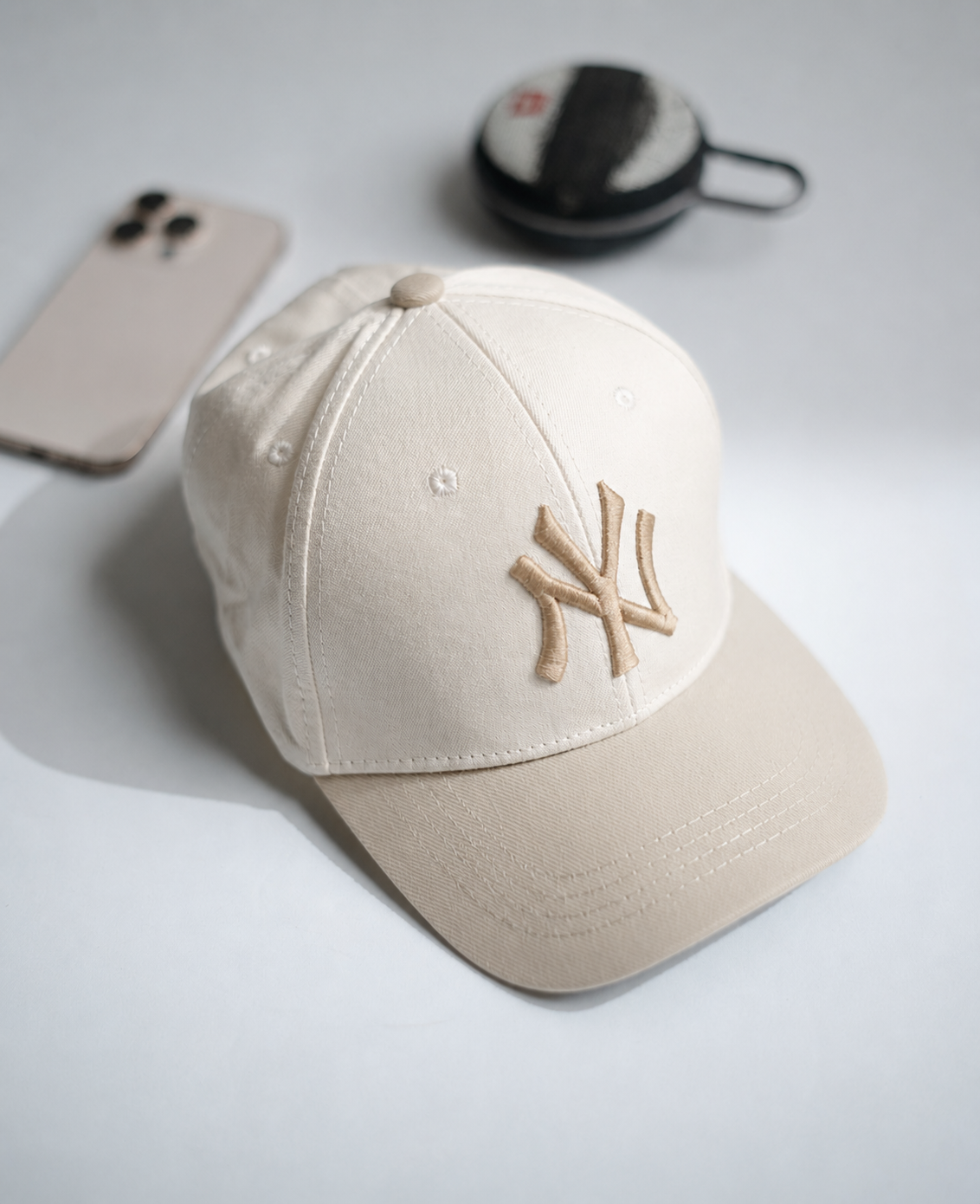 A cream-colored baseball cap with an embroidered tan New York Yankees logo on the front, styled on a light gray surface with a smartphone and a small round case in the background in Pakistan by Caps Nation