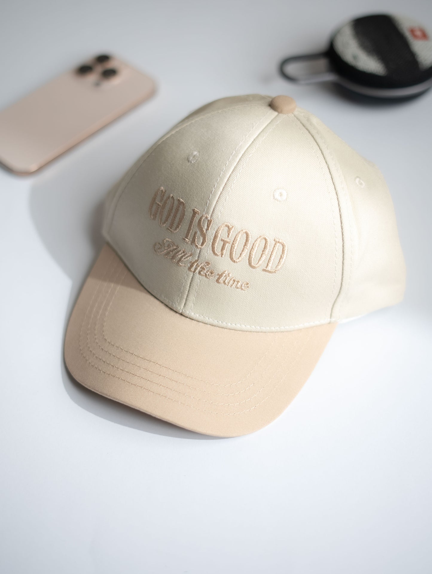 Beige cap with 'God is Good' text on a light gray surface with phone and wallet in the background in Pakistan by Caps Nation
