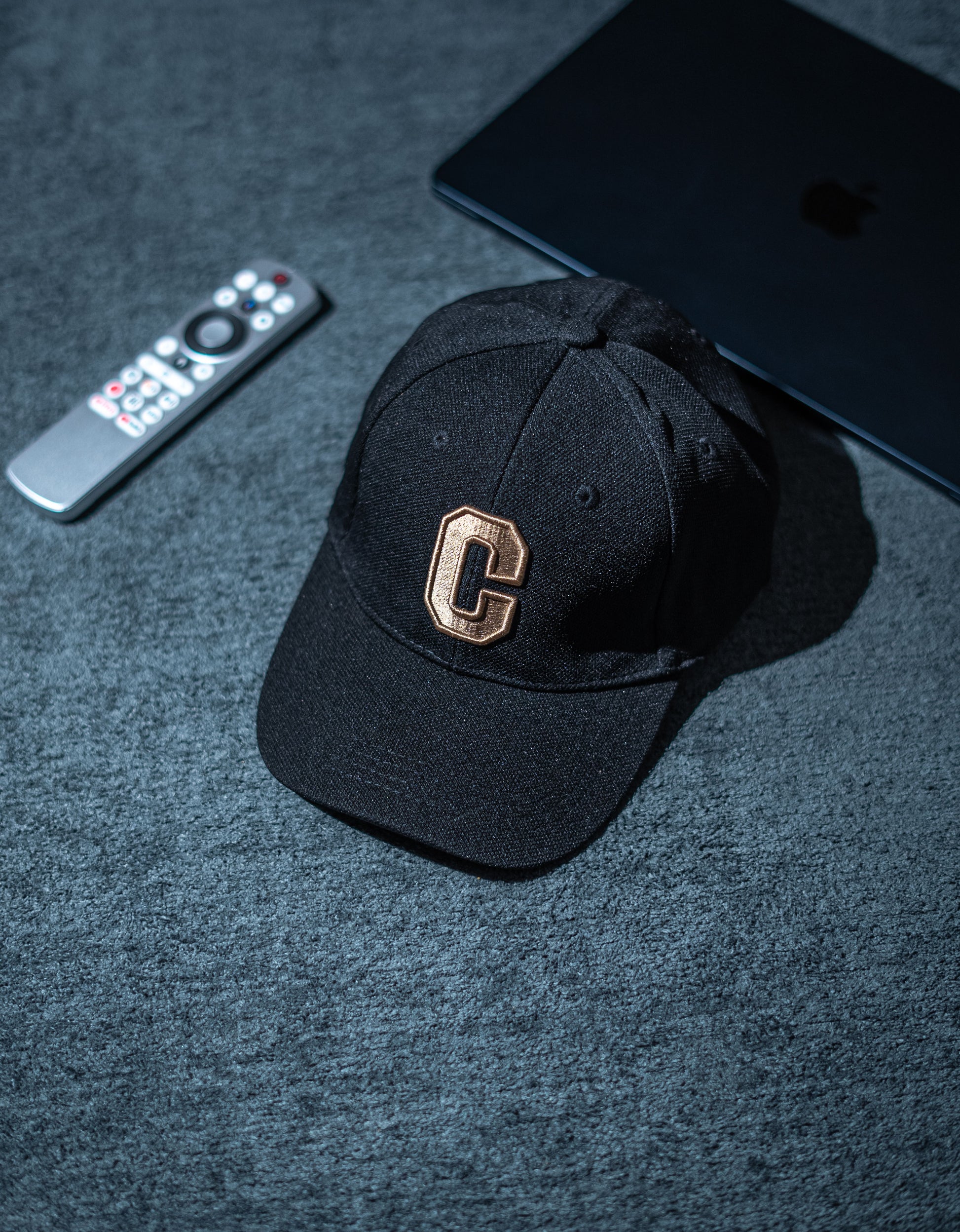 Black cap with a gold logo on a dark surface next to a remote control and tablet in Pakistan by Caps Nation