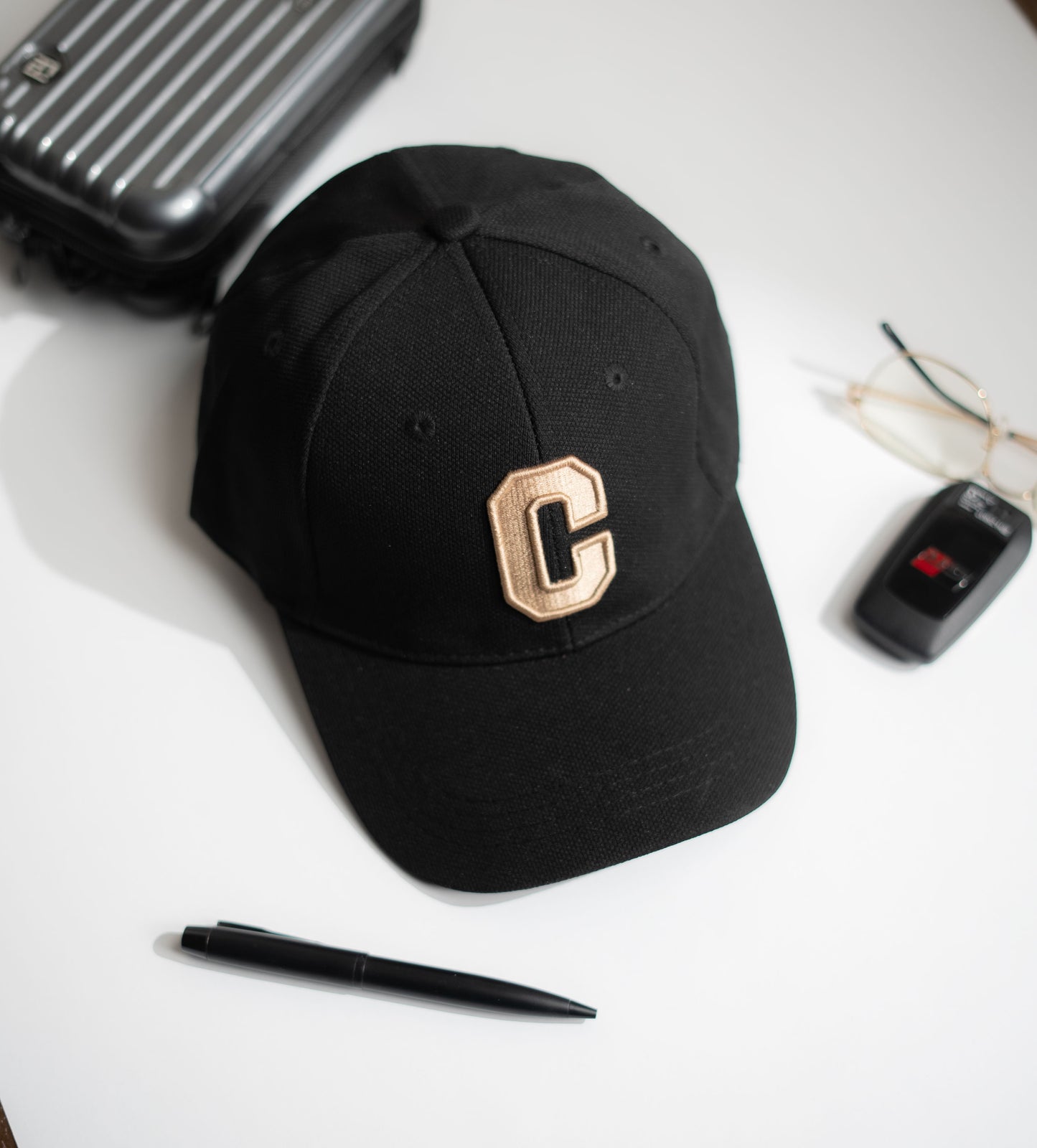 Black cap with a gold 'C' on a white surface with a suitcase, pen, and glasses in Pakistan by Caps Nation