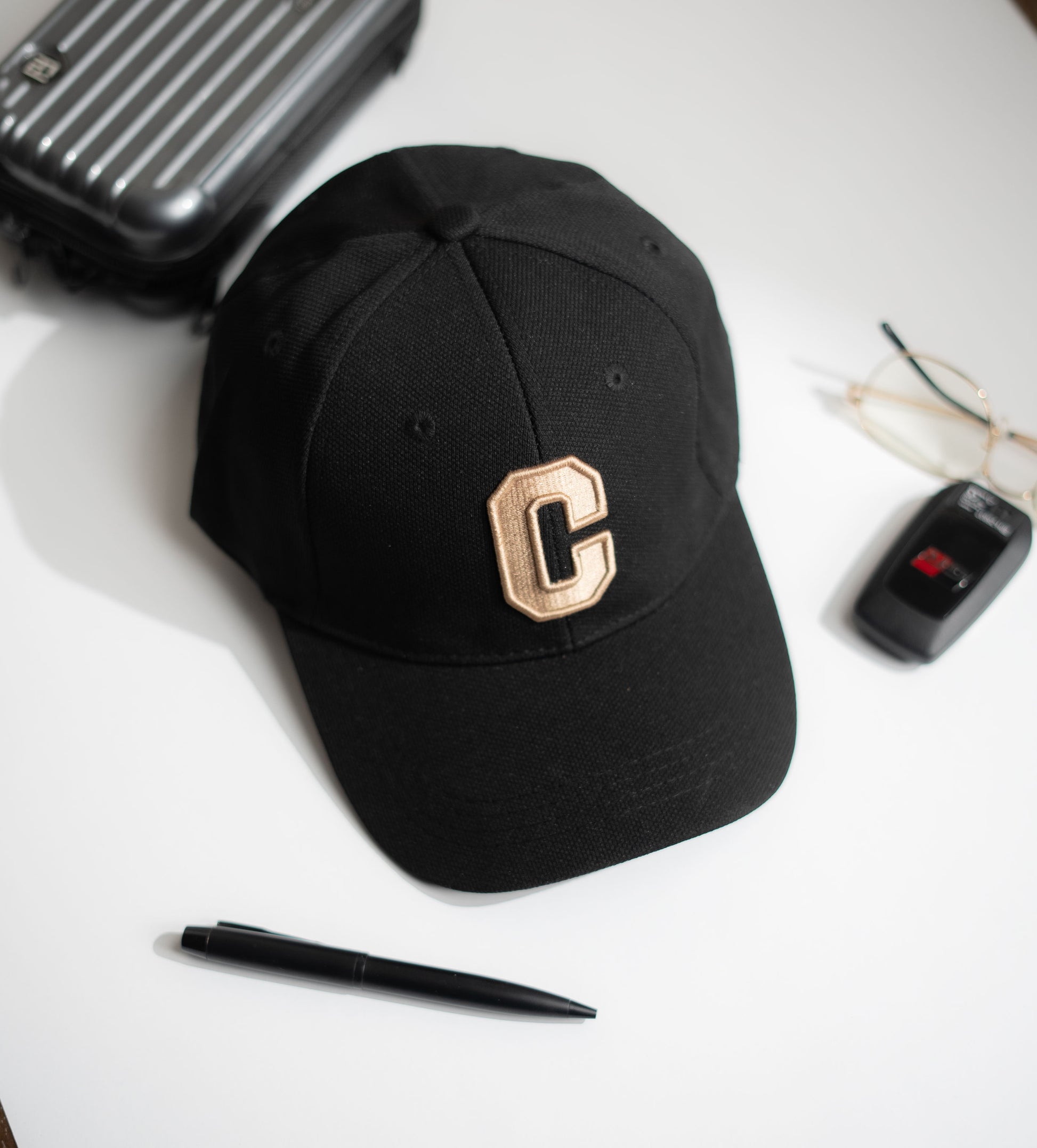 Black cap with a gold 'C' on a white surface with a suitcase, pen, and glasses in Pakistan by Caps Nation