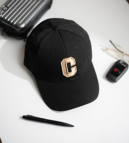 Black cap with a gold 'C' on a white surface with a suitcase, pen, and glasses in Pakistan by Caps Nation