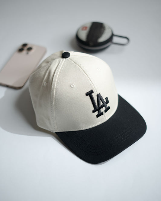 White and black cap with LA logo on a light gray surface, with phone and wallet in the background in Pakistan by Caps Nation