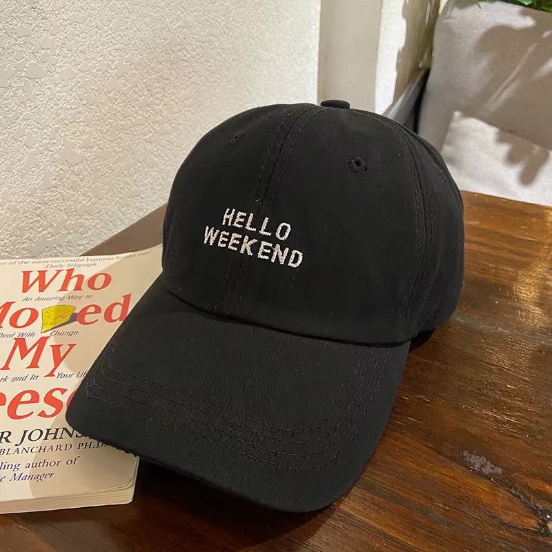 Black cap with 'Hello Weekend' text on a wooden surface next to a book in Pakistan by Caps Nation