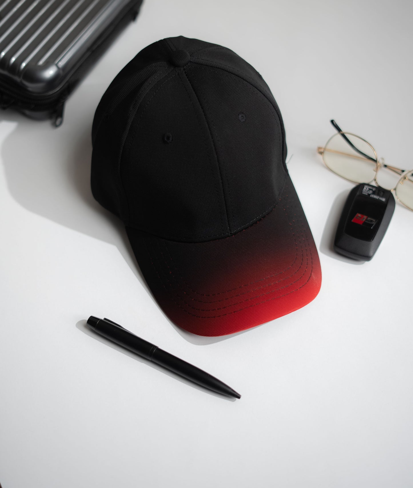 Black and red gradient cap on a white surface with a pen, glasses, and suitcase in Pakistan by Caps Nation
