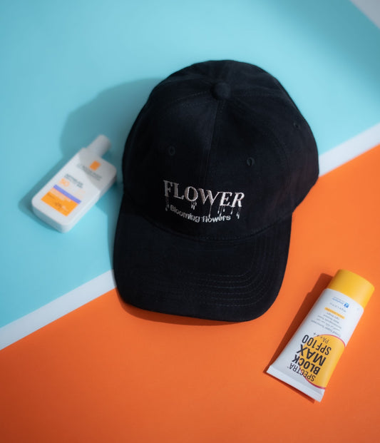 A black baseball cap with 'FLOWER blooming flowers' embroidered on the front, styled on a split blue and orange background with sunscreen bottles next to it in Pakistan by Caps Nation