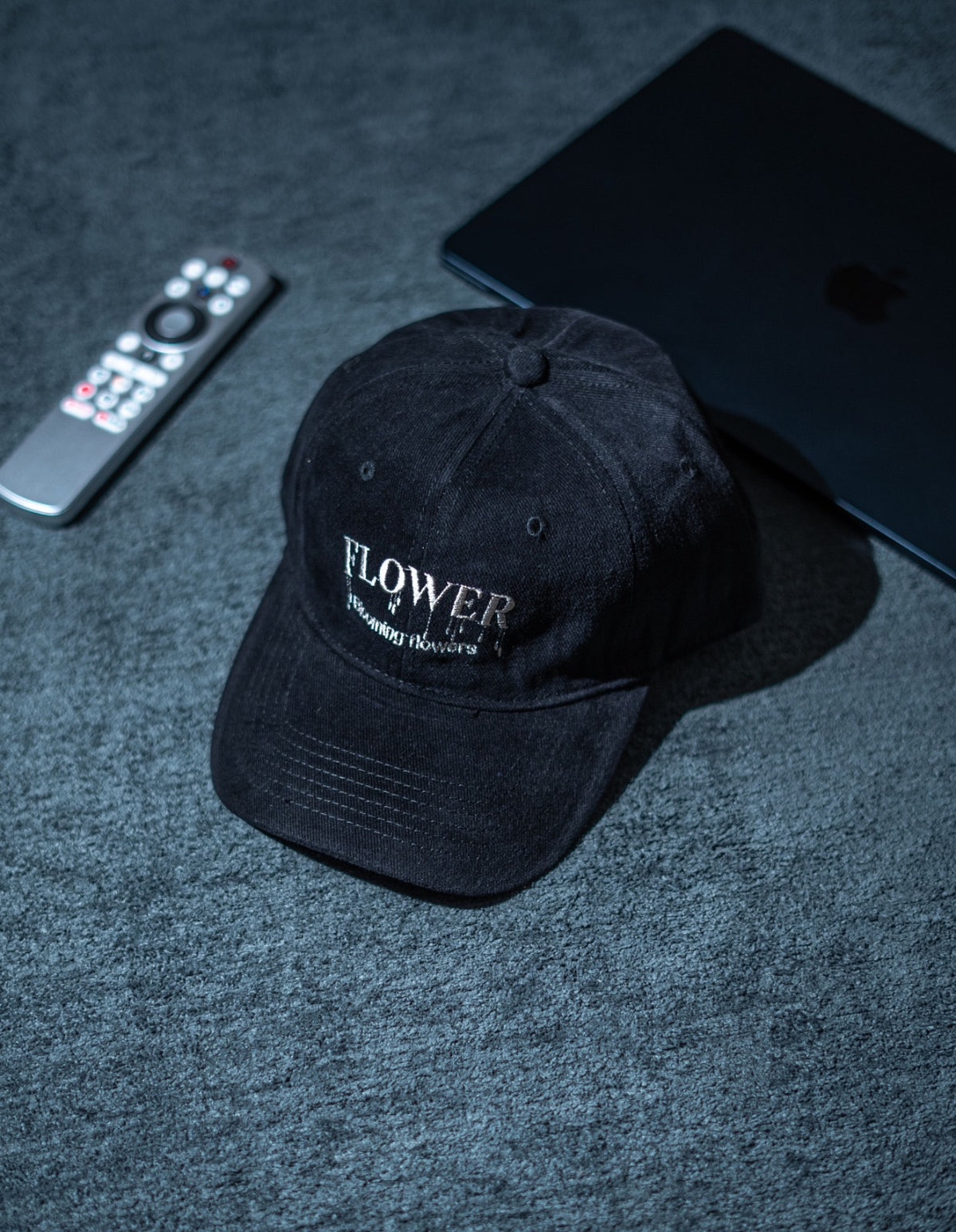 A black baseball cap with white embroidered text reading 'FLOWER' and 'The Flower Merchant' resting on a textured gray surface, styled with a remote control and a closed laptop in the background in Pakistan by Caps Nation