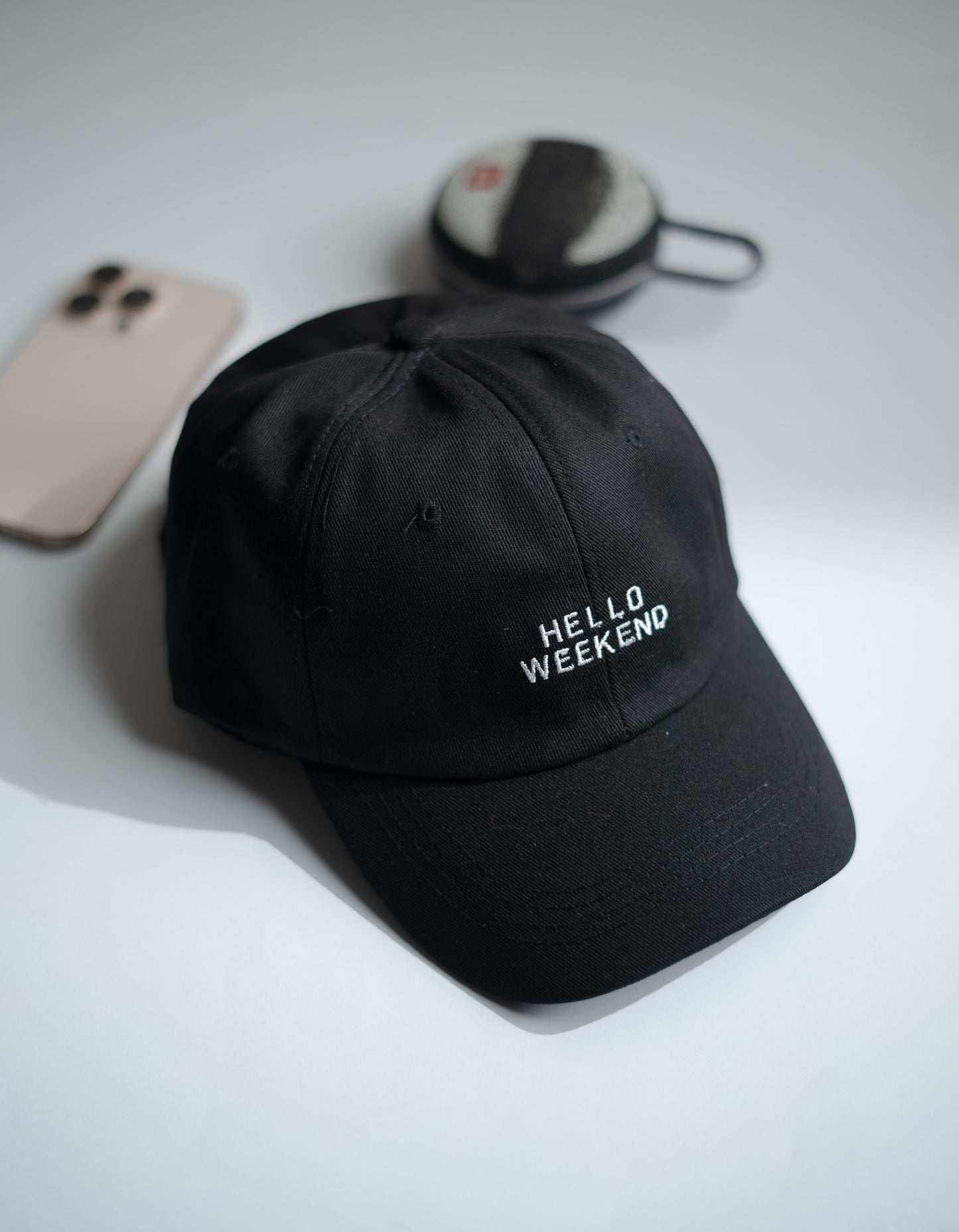 Black cap with 'Hello Weekend' text on a light gray surface with phone and earbuds in Pakistan by Caps Nation