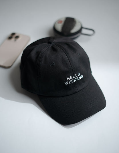 Black cap with 'Hello Weekend' text on a light gray surface with phone and earbuds in Pakistan by Caps Nation