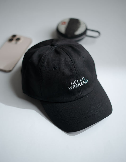 Black cap with 'Hello Weekend' text on a light gray surface with phone and earbuds in Pakistan by Caps Nation