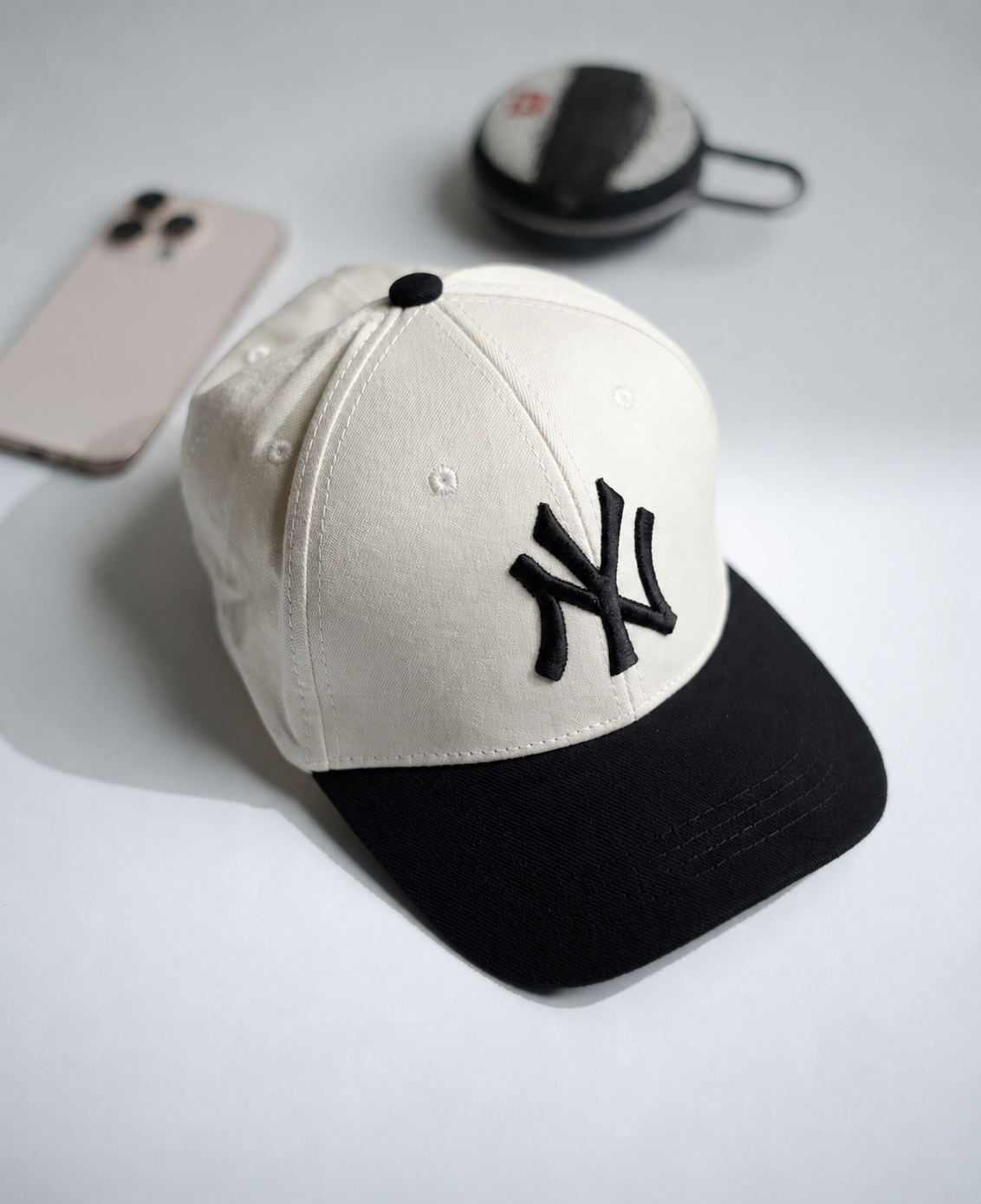 A baseball cap with an off-white crown and a black brim, featuring a black embroidered New York Yankees logo on the front. The cap is styled on a light gray surface with a smartphone and a small round case in the background in Pakistan by Caps Nation