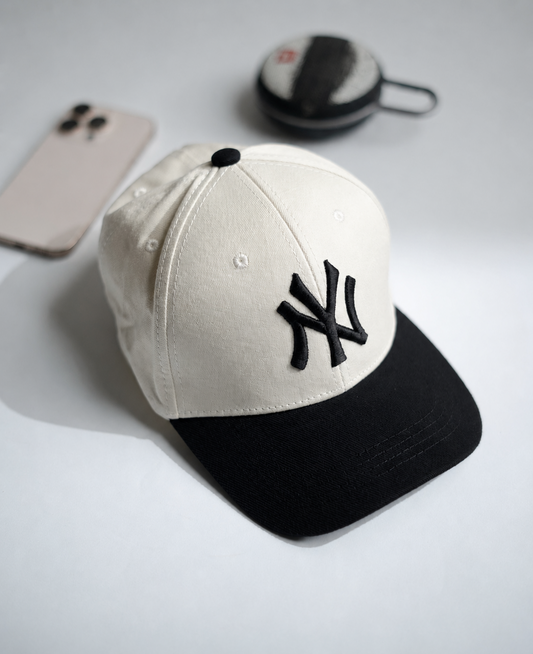 A baseball cap with an off-white crown and a black brim, featuring a black embroidered New York Yankees logo on the front. The cap is styled on a light gray surface with a smartphone and a small round case in the background in Pakistan by Caps Nation