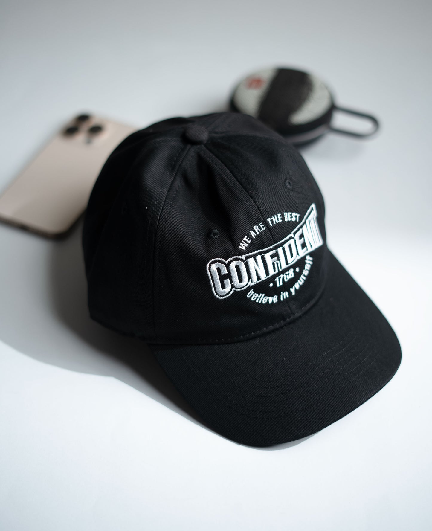 Black cap with white text on a light gray surface with a phone and sunglasses in Pakistan by Caps Nation