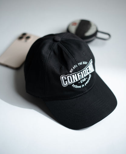 Black cap with white text on a light gray surface with a phone and sunglasses in Pakistan by Caps Nation