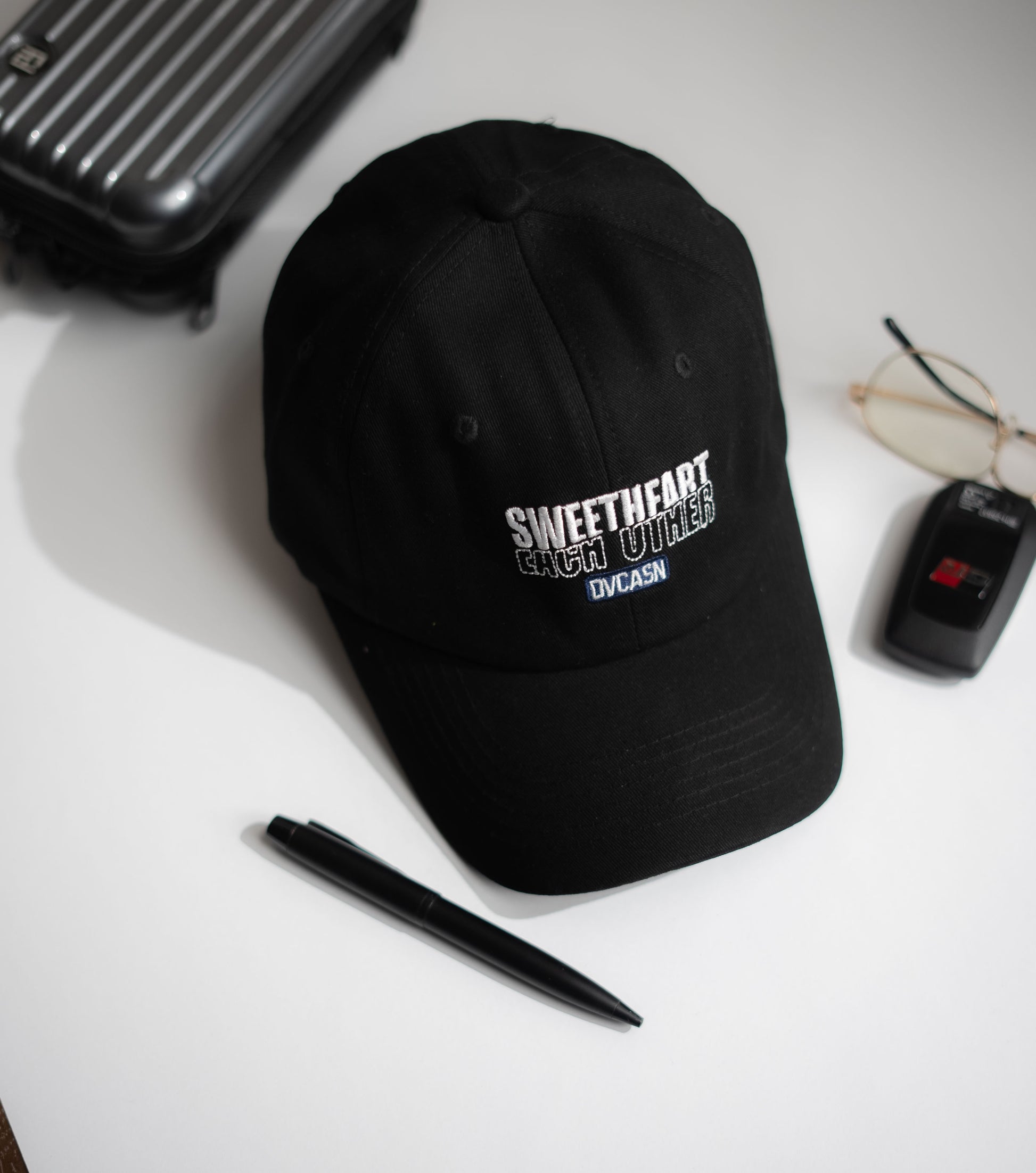 Black cap with text on a white surface with a pen, glasses, and suitcase in Pakistan by Caps Nation
