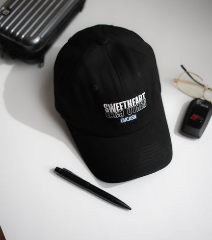 Black cap with text on a white surface with a pen, glasses, and suitcase in Pakistan by Caps Nation