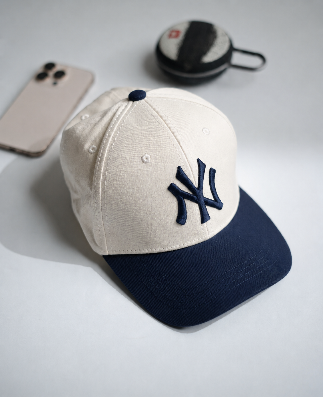 A New York Yankees baseball cap with an off-white crown and a navy blue visor, styled on a light gray surface with a smartphone and a small round case in the background in Pakistan by Caps Nation