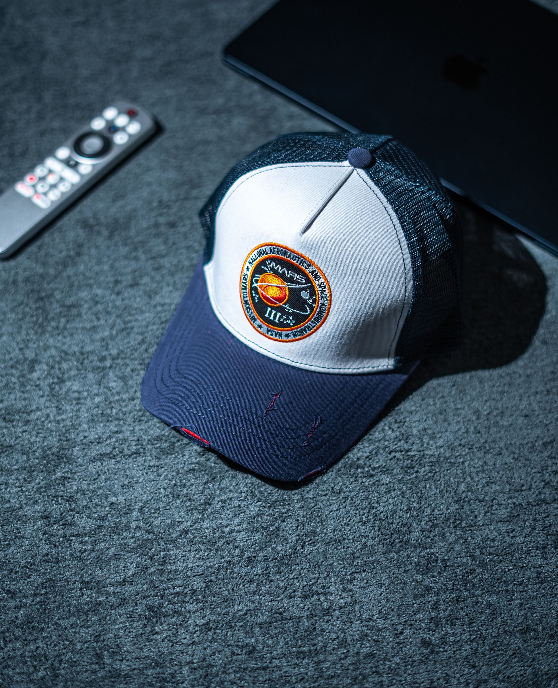 Blue and white cap with a logo on a dark surface next to a remote control and tablet in Pakistan by Caps Nation