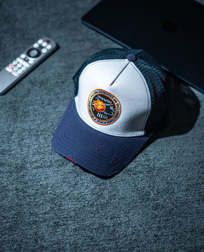 Blue and white cap with a logo on a dark surface next to a remote control and tablet in Pakistan by Caps Nation