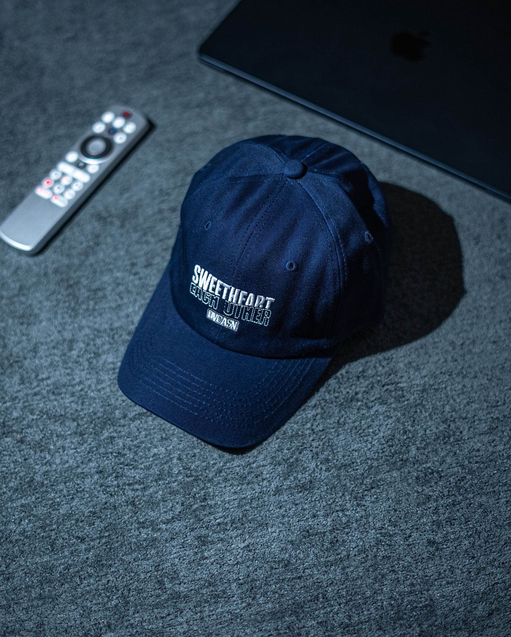 Navy blue cap with branding on a dark textured surface with remote control and tablet in Pakistan by Caps Nation