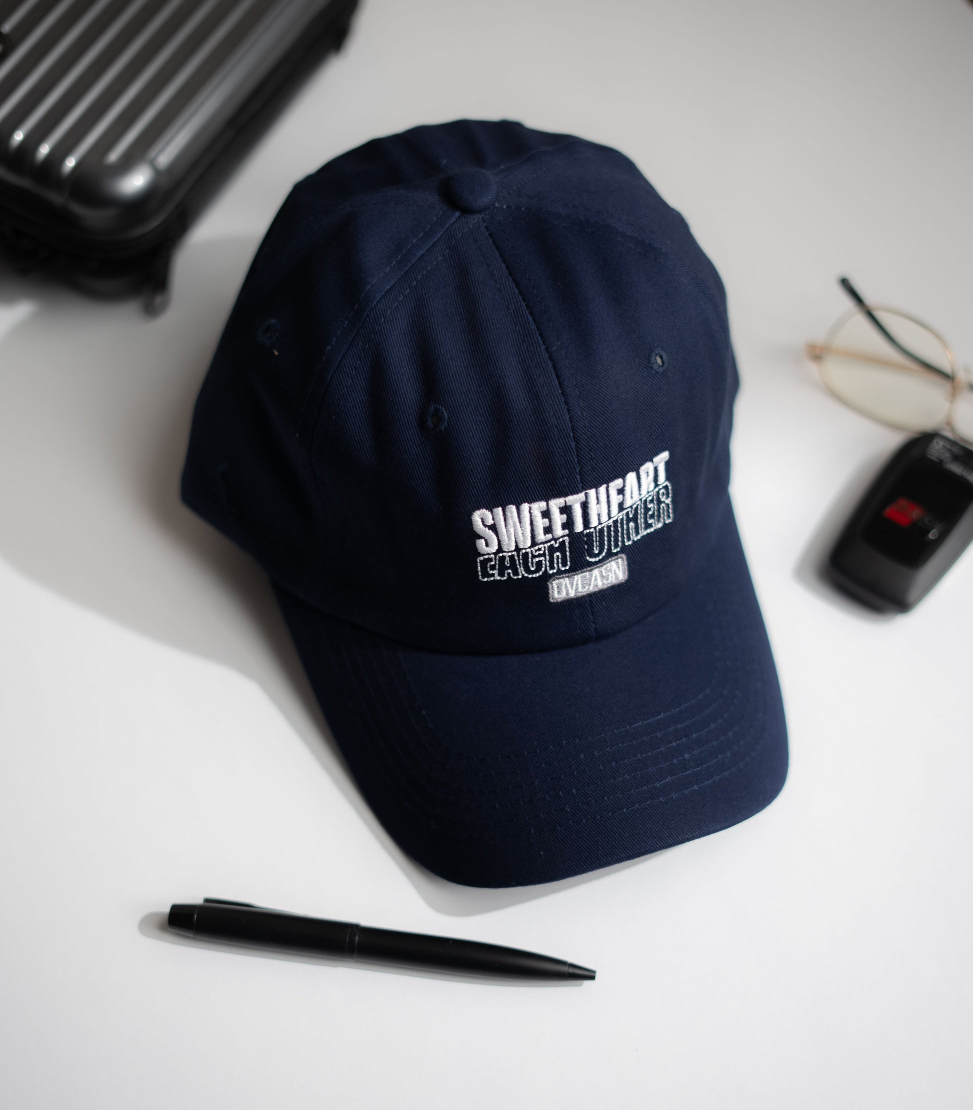Navy blue cap with text on a white surface with a pen, glasses, and black case in Pakistan by Caps Nation