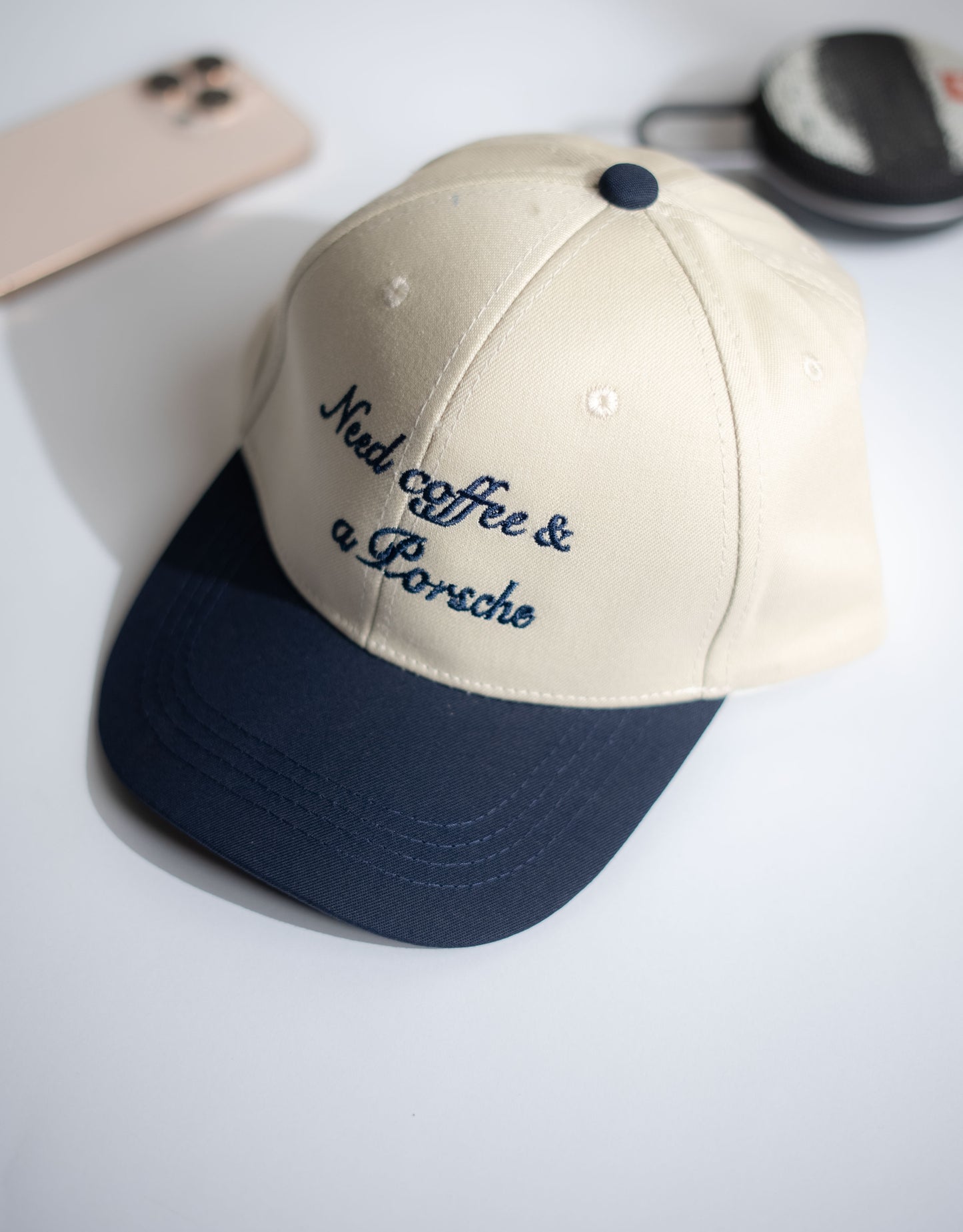 Beige and navy cap with 'Need coffee & a Porsche' text on a white surface in Pakistan by Caps Nation
