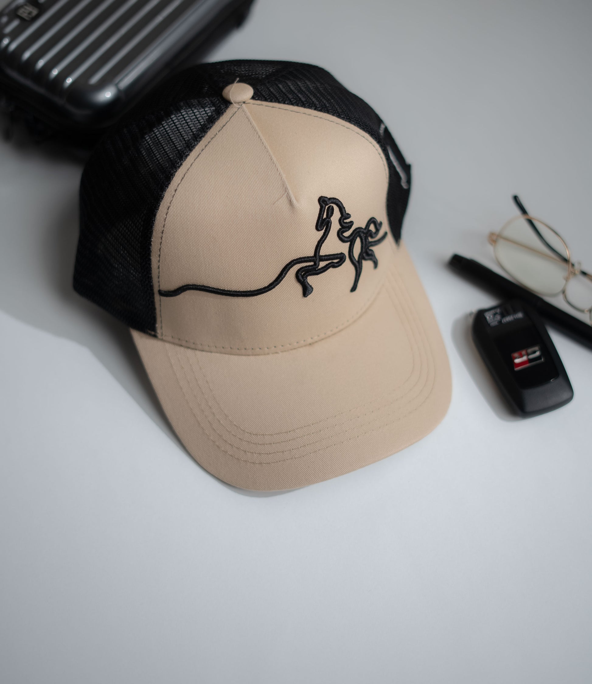 Beige cap with black horse design on a white surface with phone, glasses, and car key in Pakistan by Caps Nation