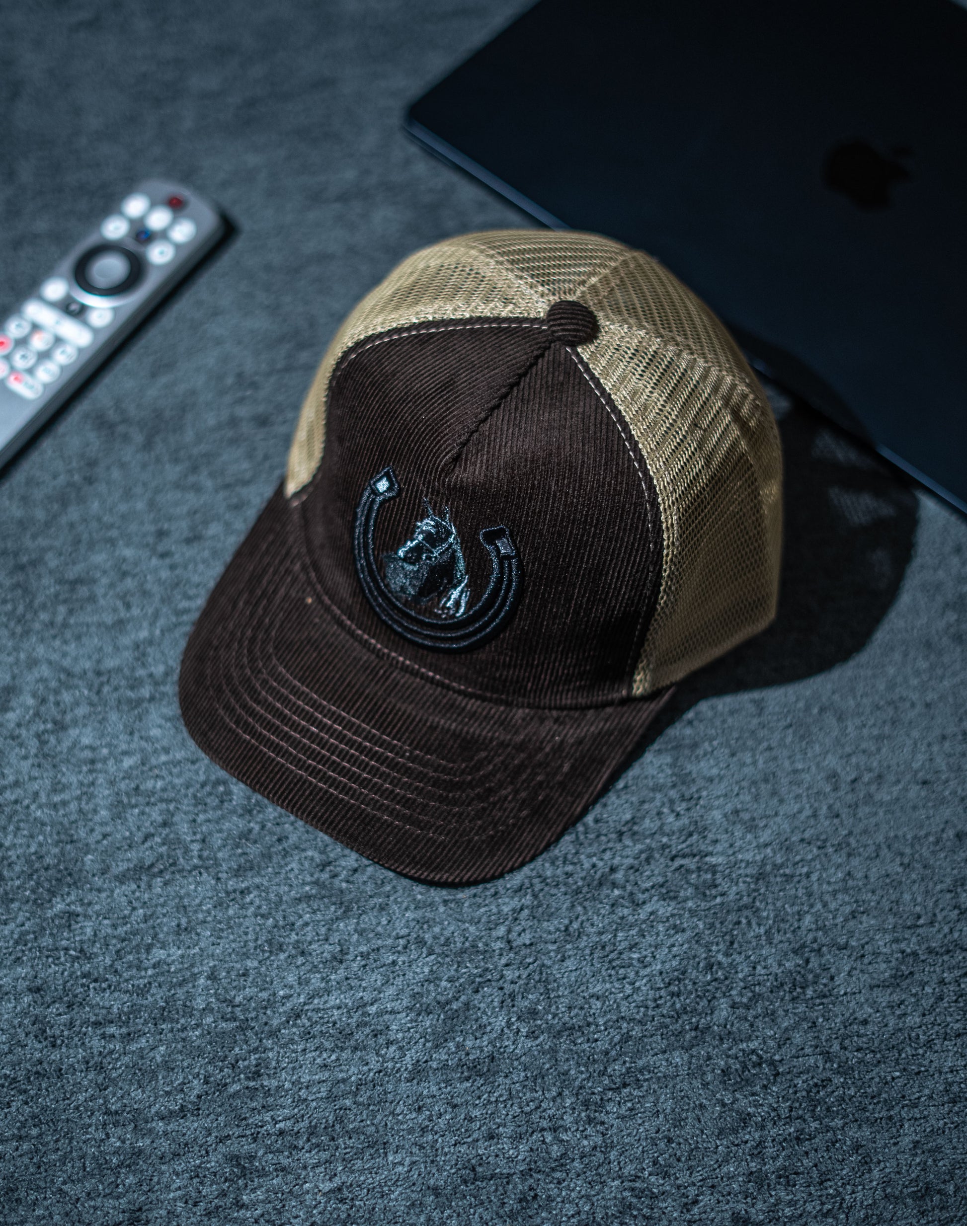 Brown and beige cap with a logo on a dark textured surface with remote control and black device in Pakistan by Caps Nation