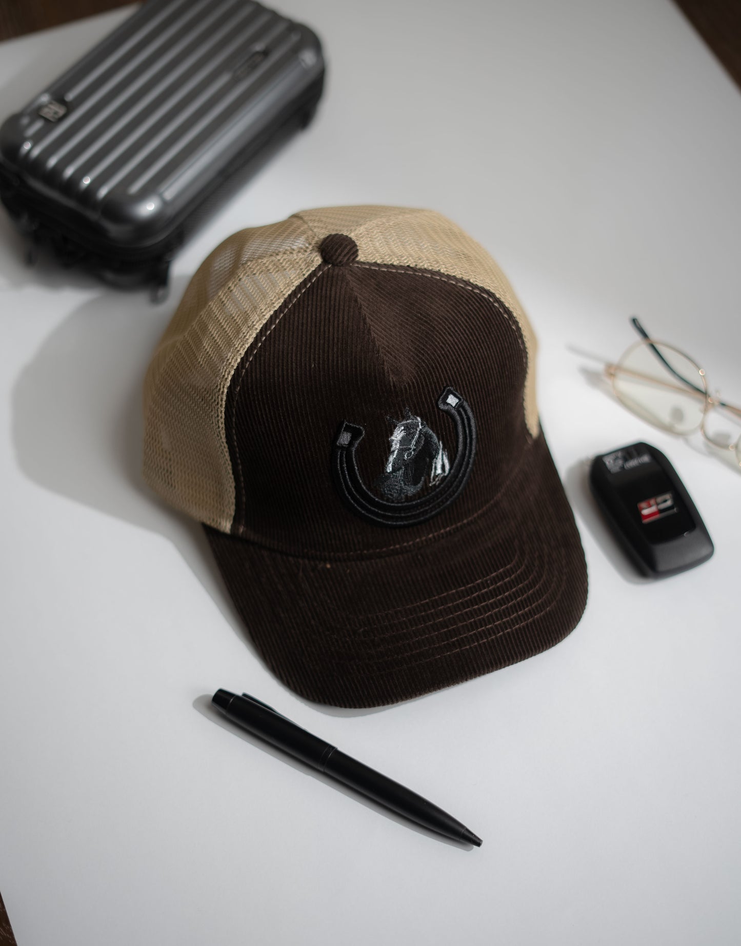 Brown and beige cap with a logo on a white surface, next to a pen and small black device in Pakistan by Caps Nation