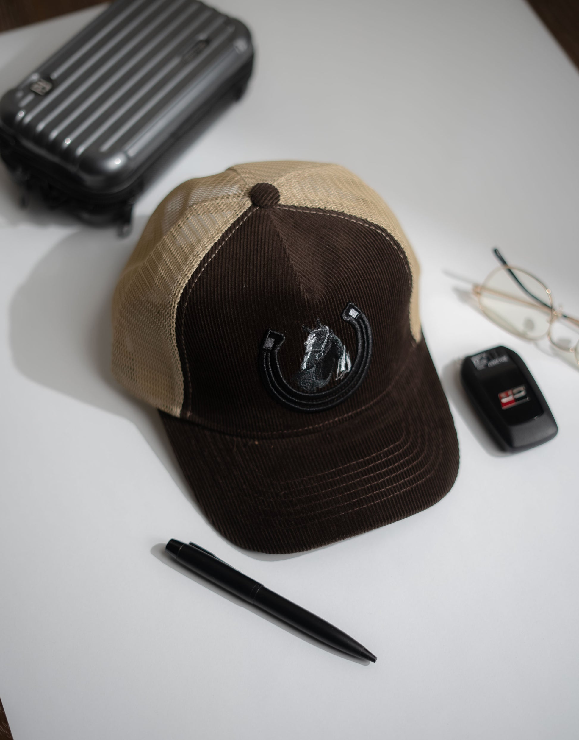 Brown and beige cap with a logo on a white surface, next to a pen and small black device in Pakistan by Caps Nation