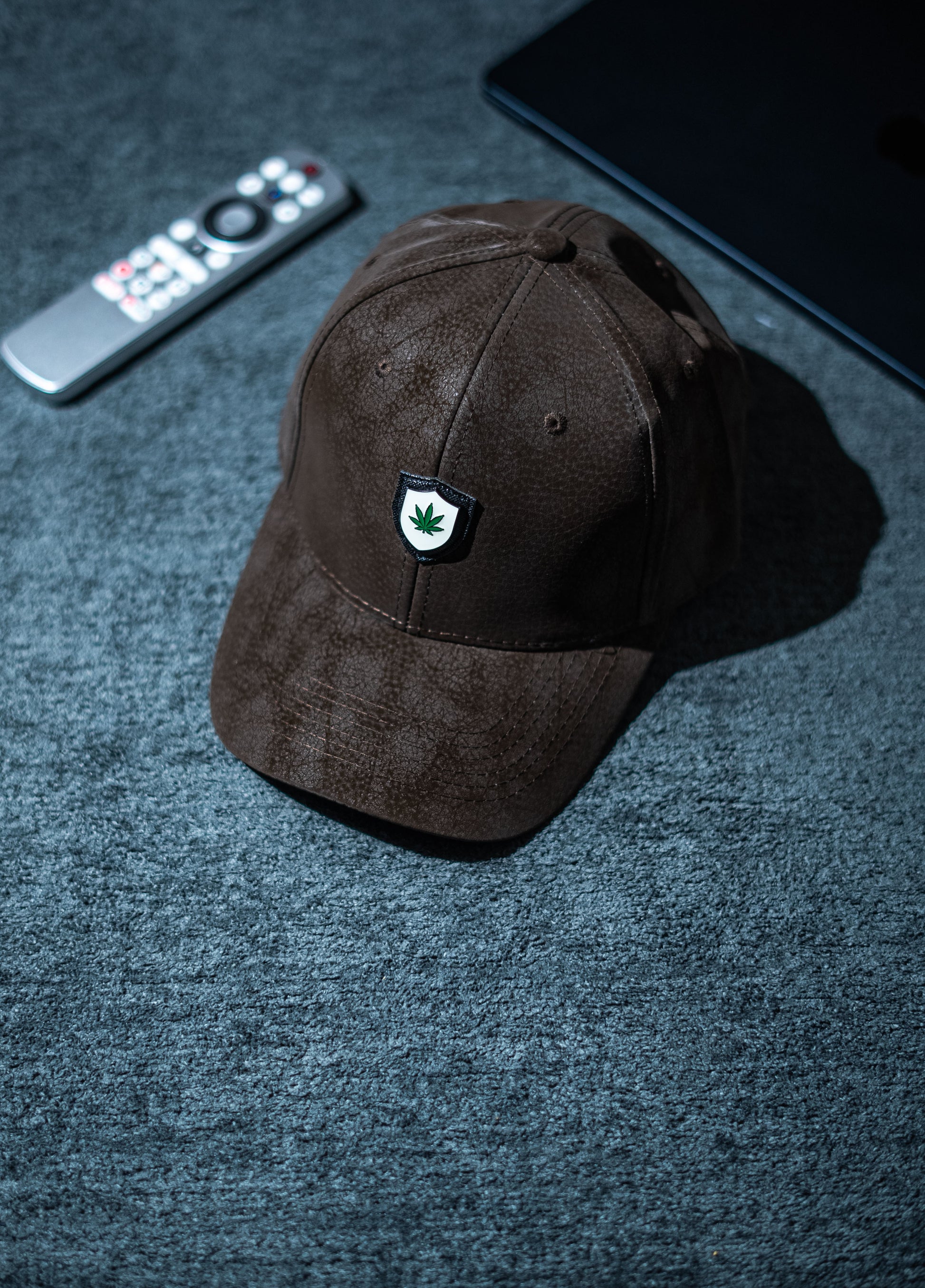 Brown cap with a logo on a dark textured surface with a remote control and phone in Pakistan by Caps Nation