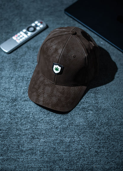Brown cap with a logo on a dark textured surface with a remote control and phone in Pakistan by Caps Nation