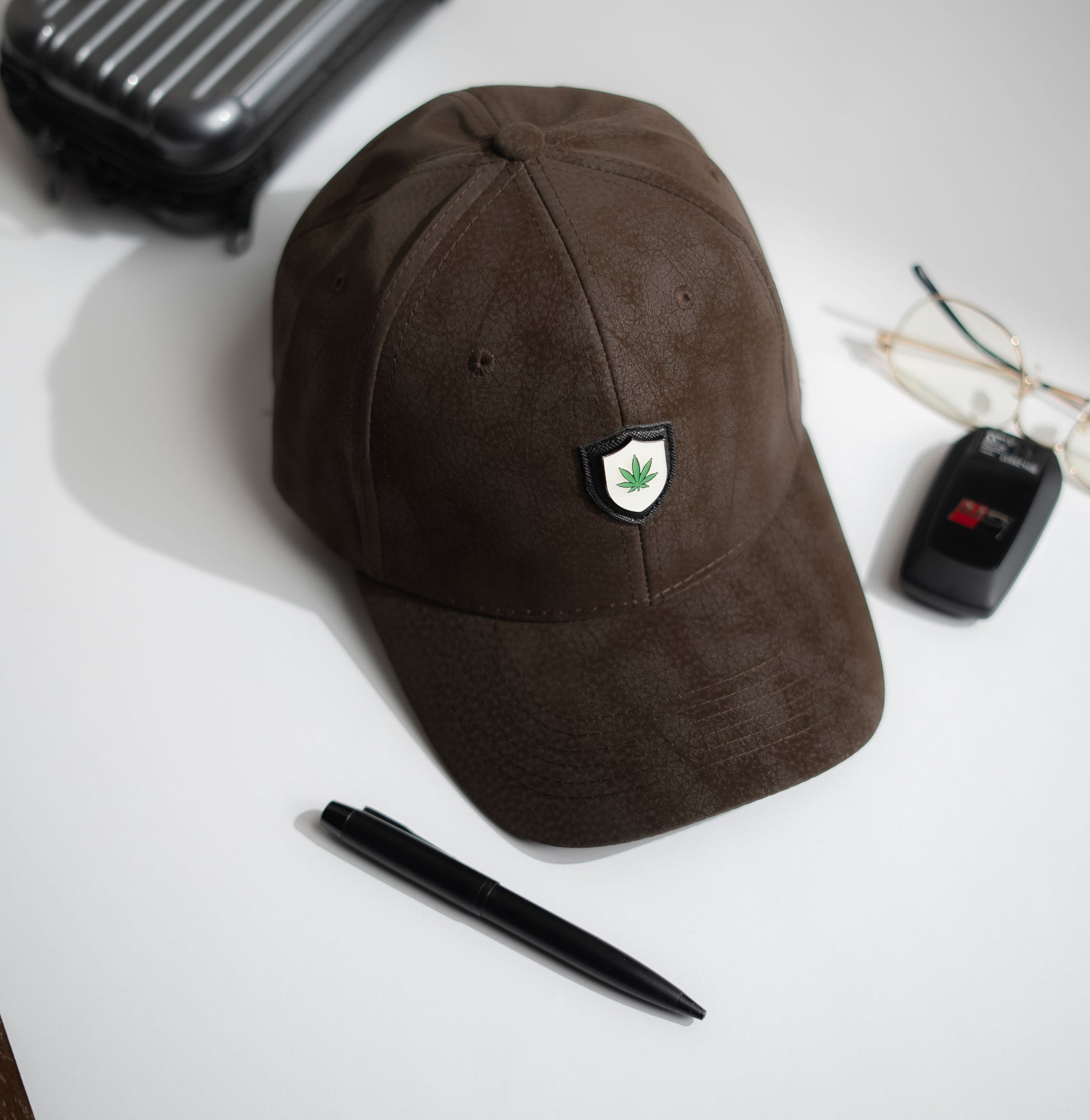 Brown cap with a logo on a white surface with a pen, car key, and suitcase in Pakistan by Caps Nation