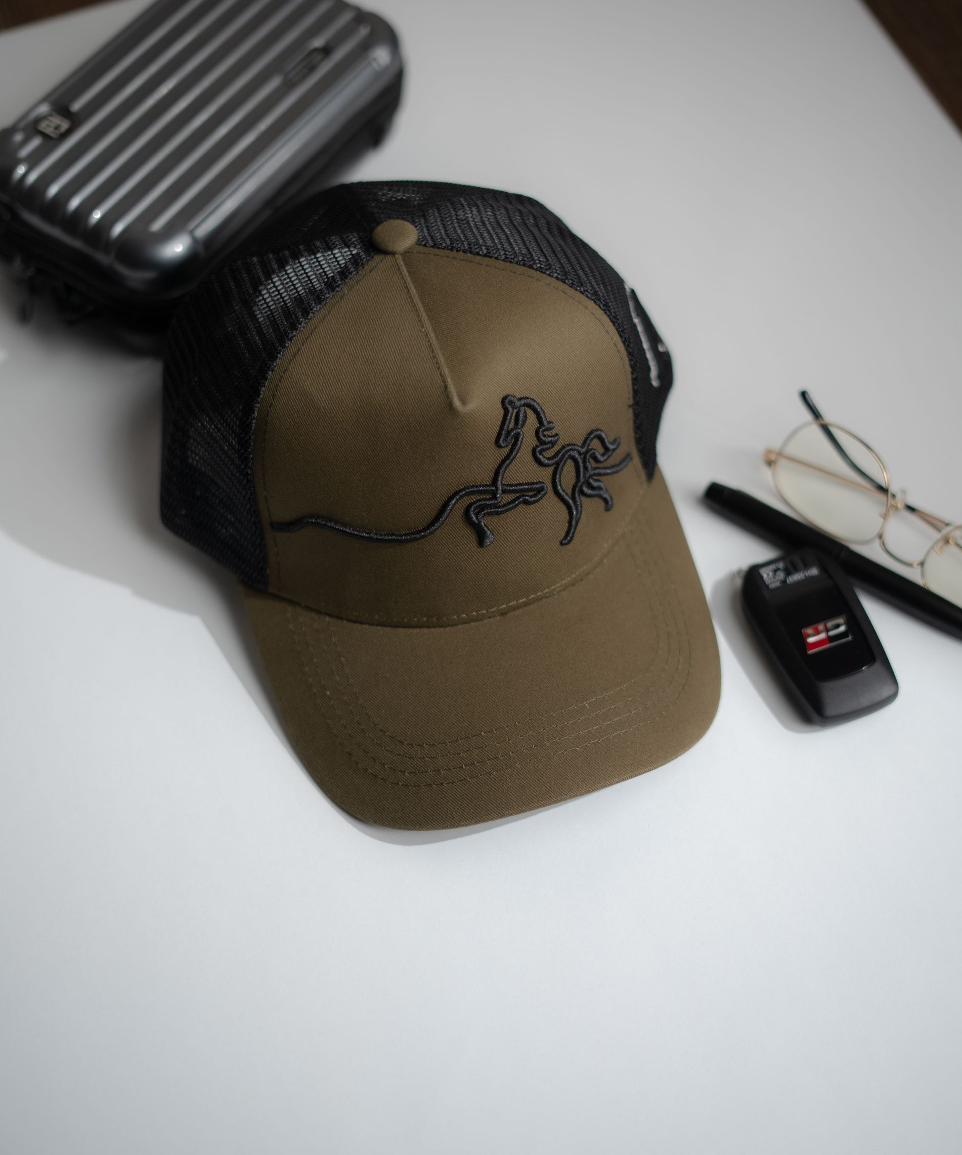 Brown cap with a horse logo on a white surface with a suitcase, glasses, and car key visible in Pakistan by Caps Nation