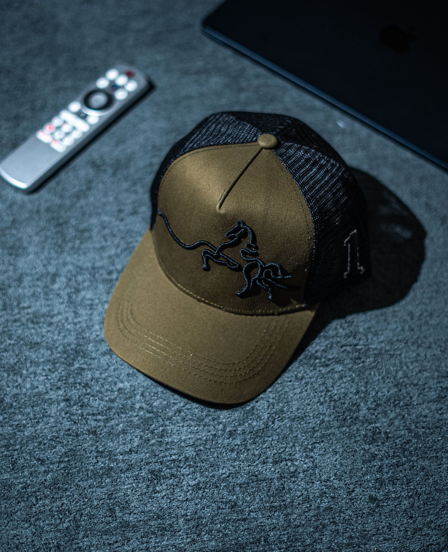 Brown cap with black logo on a dark textured surface with remote control and phone in Pakistan by Caps Nation