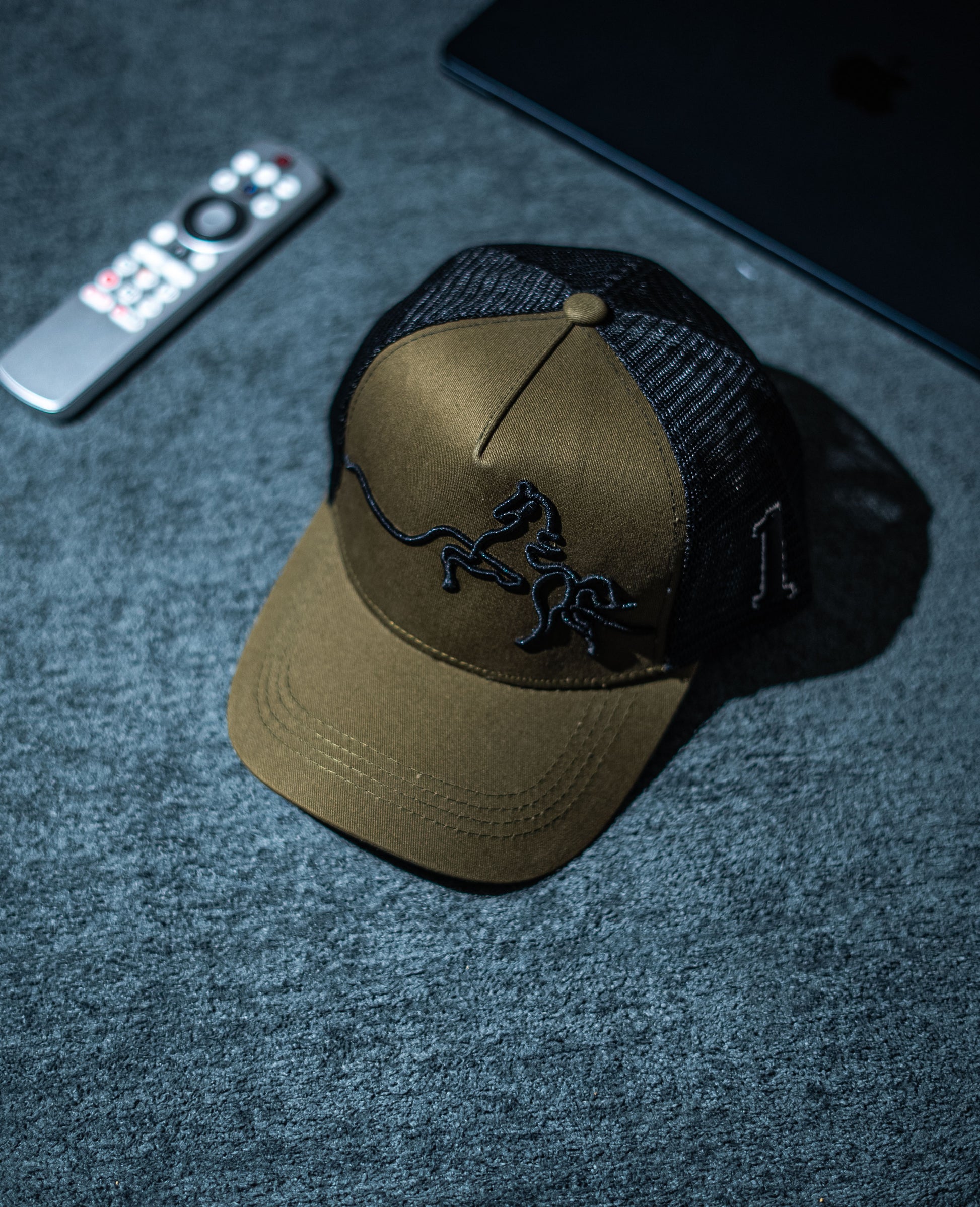 Brown cap with black logo on a dark textured surface with remote control and phone in Pakistan by Caps Nation