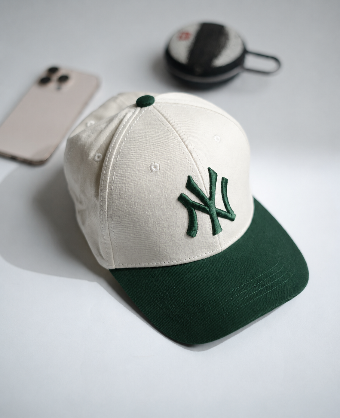 A baseball cap with an off-white crown and a dark green visor, featuring an embroidered green New York Yankees logo on the front. The cap is styled on a light gray surface with a smartphone and a small round case in the background in Pakistan by Caps Nation