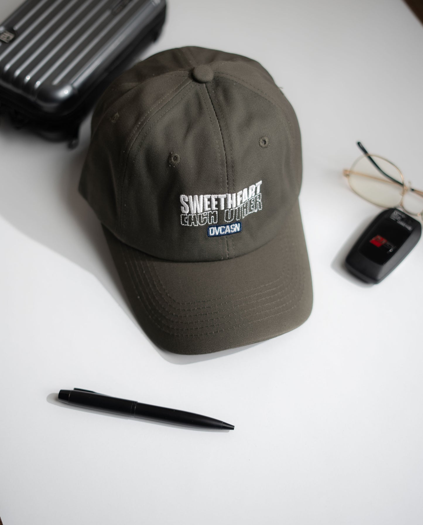 Dark gray cap with text on a white surface with a pen, glasses, and suitcase in Pakistan by Caps Nation