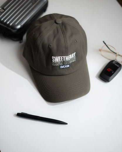 Dark gray cap with text on a white surface with a pen, glasses, and suitcase in Pakistan by Caps Nation