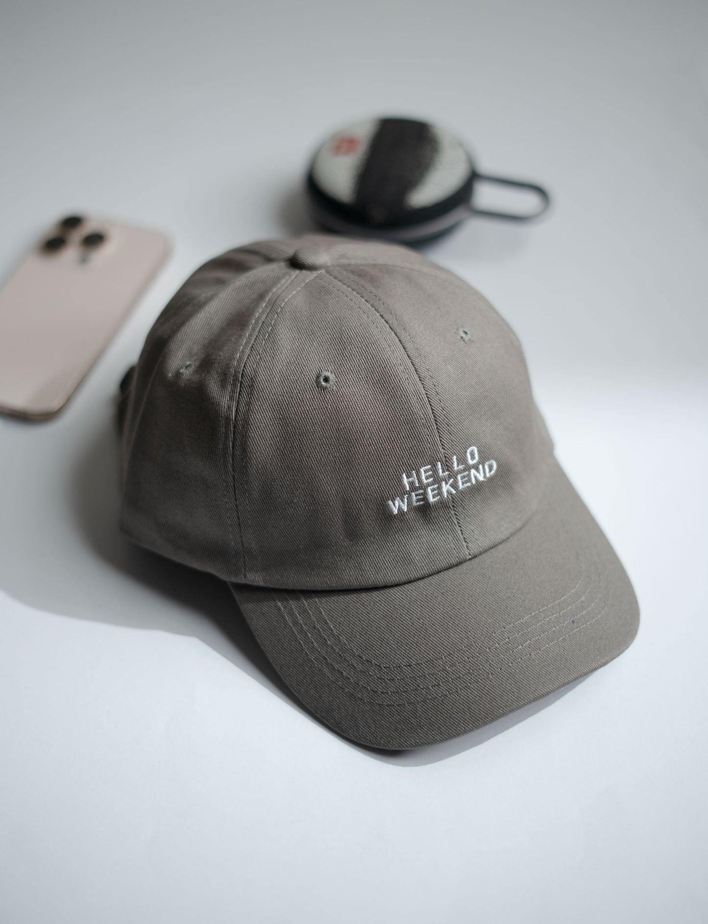 Gray cap with 'Hello Weekend' text on a light gray surface with phone and pouch in Pakistan by Caps Nation