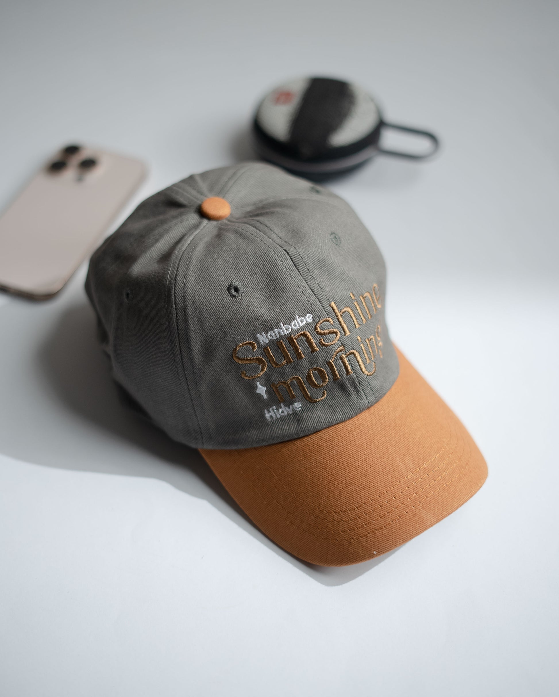 Gray cap with brown visor and embroidered text on a light gray surface with phone and black bag in Pakistan by Caps Nation