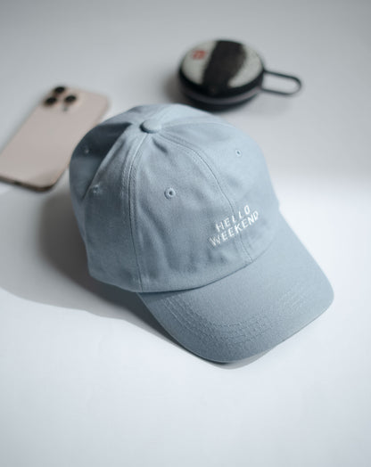 Gray cap with 'Hello Weekend' text on a light gray surface with phone and black case in Pakistan by Caps Nation