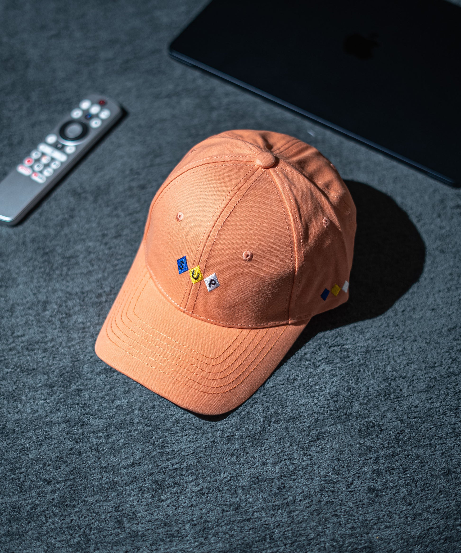 Brown cap with embroidered pins on a dark textured surface with a remote control and electronic device in Pakistan by Caps Nation