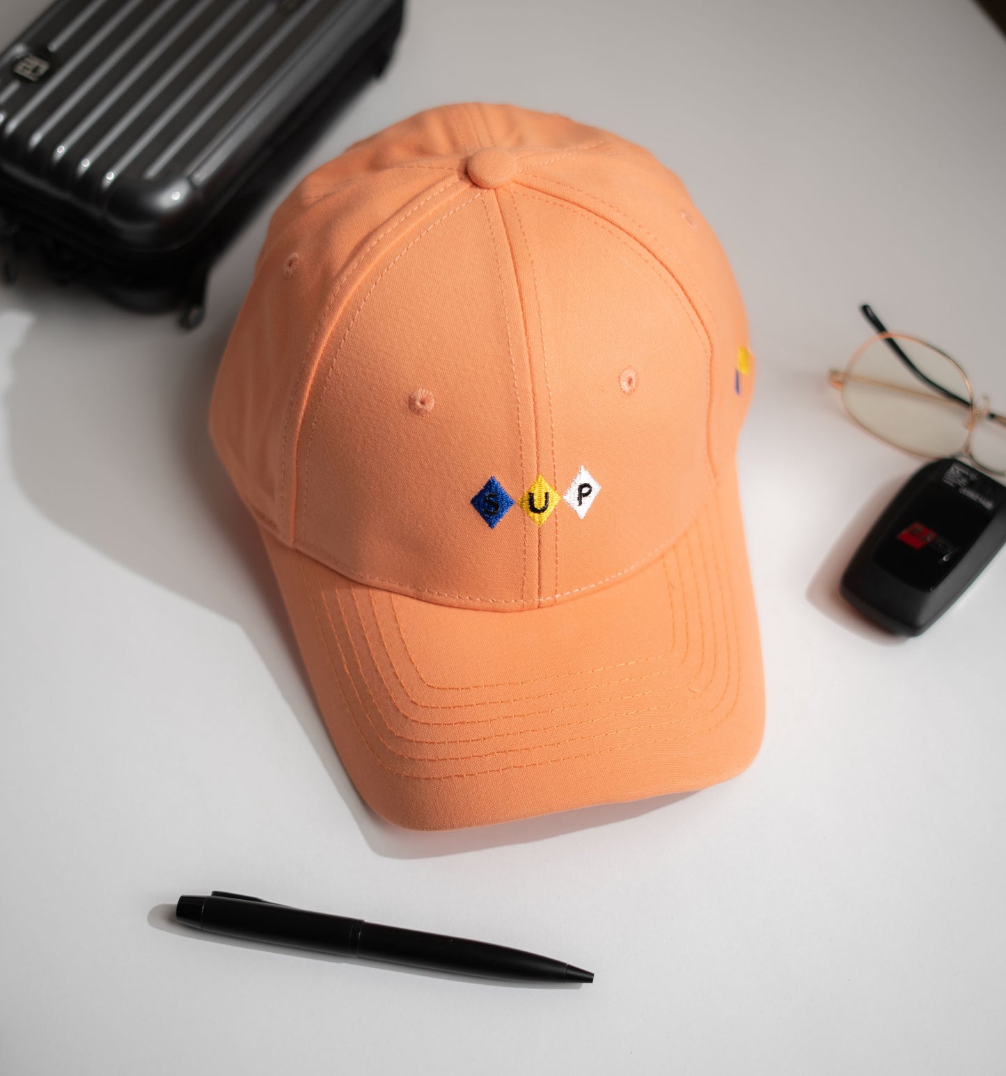 Orange cap with embroidered logos on a white surface with a suitcase, pen, and glasses in Pakistan by Caps Nation